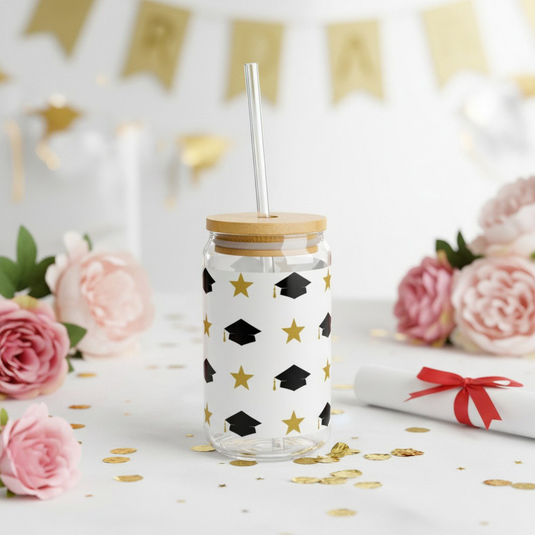 Graduation sipper glass with black grad caps and gold stars pattern on white party table with gold confetti, pink roses, diploma scroll, and gold pennant banner, Class of 2026 celebration