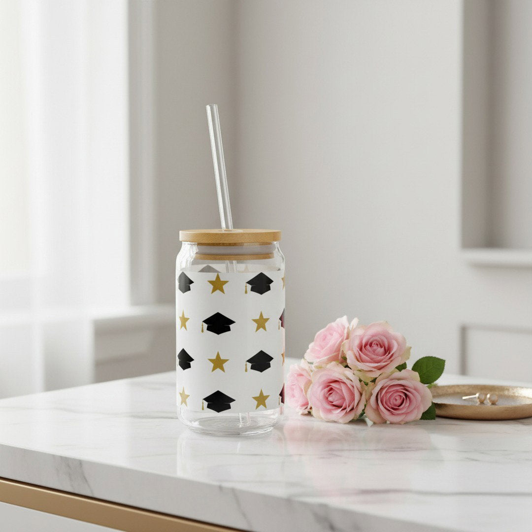 Graduation sipper glass with black grad caps and gold stars pattern on white marble surface beside pink roses and gold jewelry tray, elegant feminine graduation gift lifestyle photo