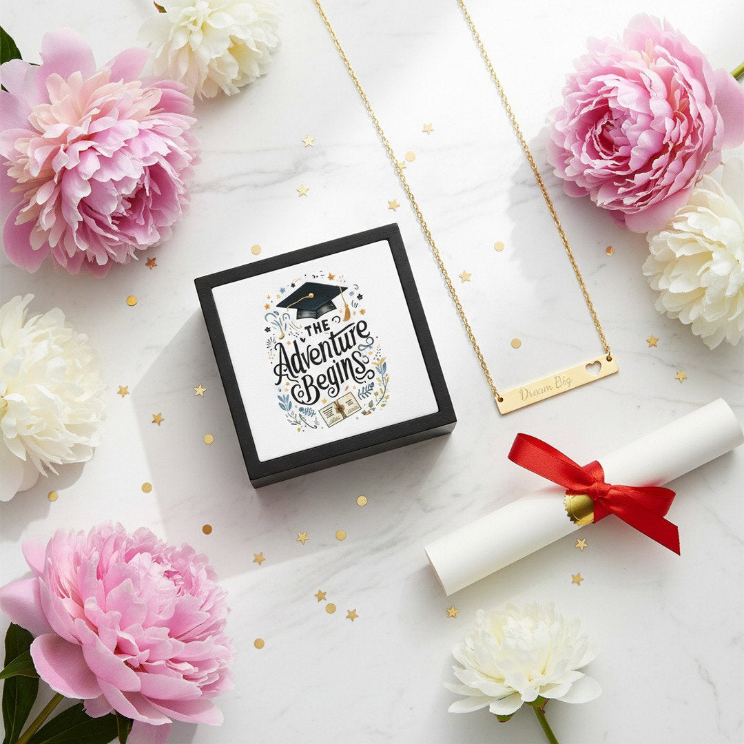 Adventure Begins graduation jewelry box with graduation cap lid design and gold Dream Big heart bar necklace on white marble surface surrounded by pink and white peonies, gold star confetti, and diploma scroll with red ribbon, graduation gift bundle flat lay