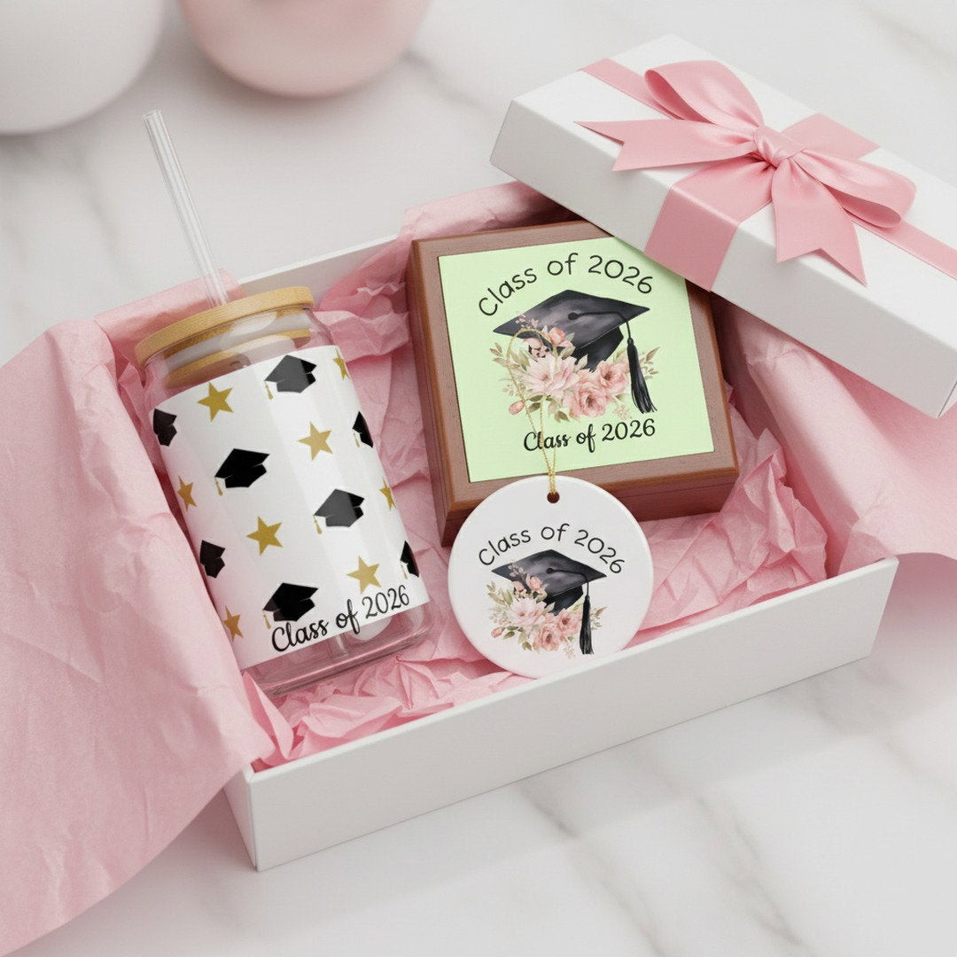 Class of 2026 graduation keepsake bundle with gold star sipper glass, floral jewelry box, and ceramic ornament nestled in open white gift box with pink tissue paper and pink satin bow, graduation gift set presentation