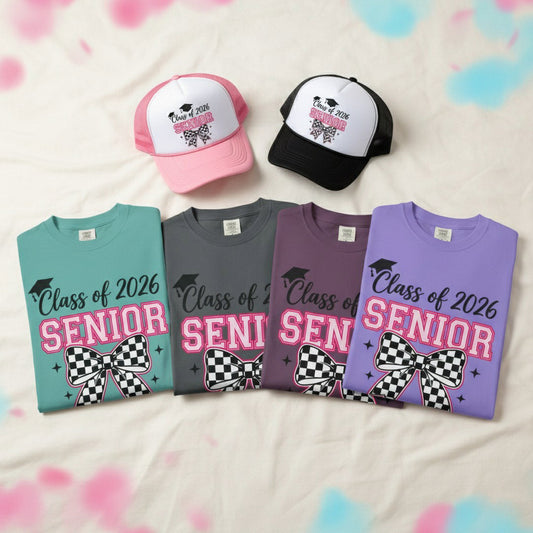  Four Class of 2026 Senior checkered bow t-shirts in seafoam, charcoal, dark berry, and violet folded in a row with pink and black Senior checkered bow trucker caps on white fabric with pink and blue confetti, graduation t-shirt and hat bundle color options