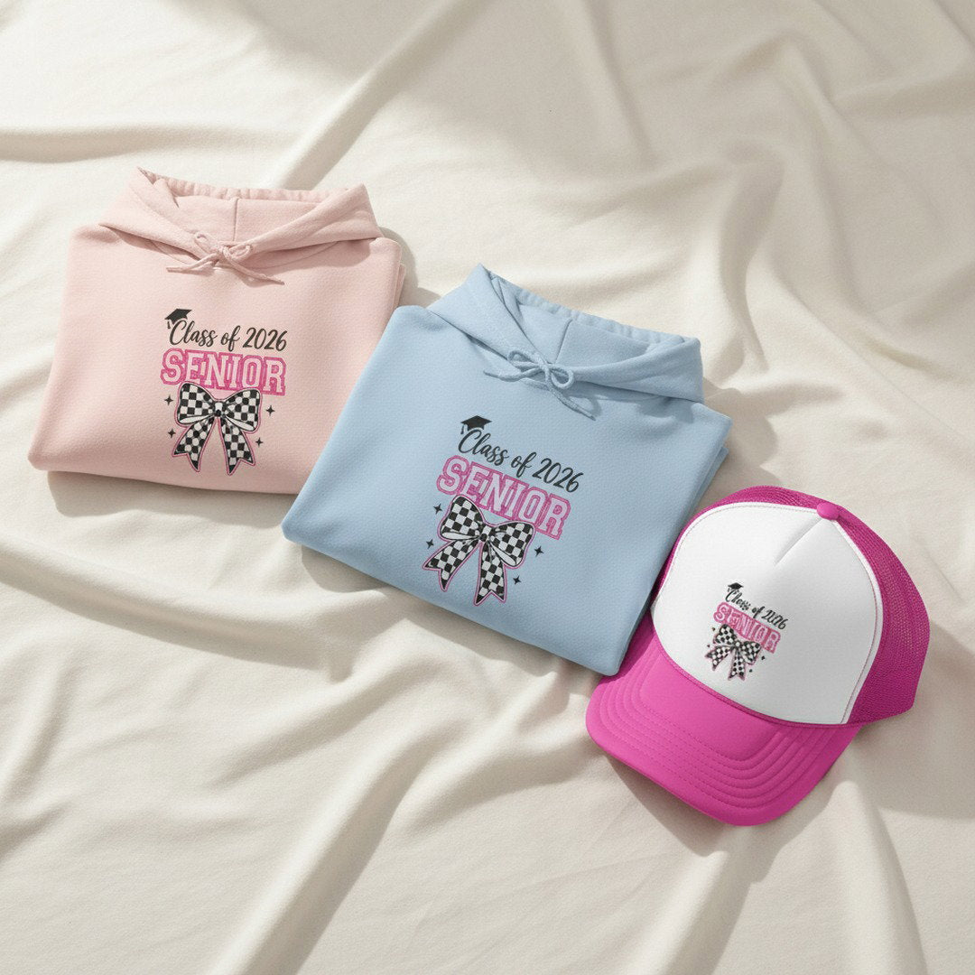 Light pink and light blue Class of 2026 Senior checkered bow hoodies folded together with pink and white Senior trucker cap on soft white fabric, graduation hoodie and hat bundle color options flat lay

